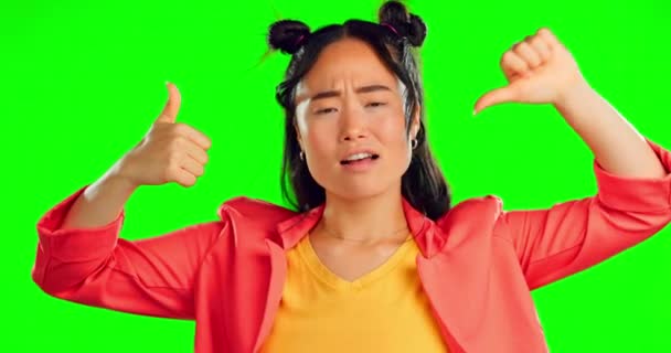 Confused, review and a woman on a green screen with a thumbs up or down isolated on a studio background. Decision, choice and portrait of an Asian girl with doubt about hand gesture feedback. - Footage, Video