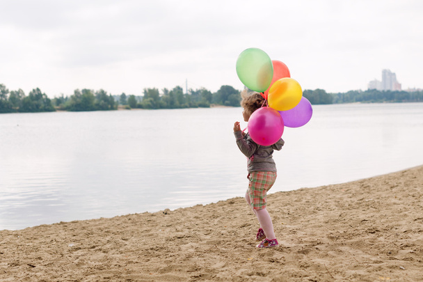 Little girl with balloons on the sandy bank of the river or lake. - Photo, Image