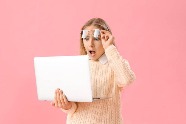 Shocked little girl in eyeglasses using laptop on pink background - Photo, Image