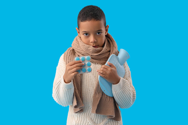 Ill African-American boy with hot water bottle and pills on blue background - Photo, Image