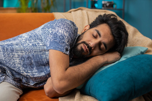 Tired indian man lying down in bed taking a rest. Carefree young hindu guy napping, falling asleep on comfortable sofa with pillows at home apartment room. Closed his eyes enjoy daytime nap alone - Foto, imagen
