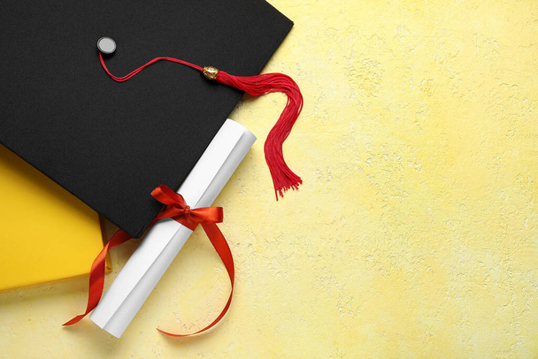 Kırmızı kurdeleli diploma, mezuniyet şapkası ve sarı masadaki kitap. - Fotoğraf, Görsel