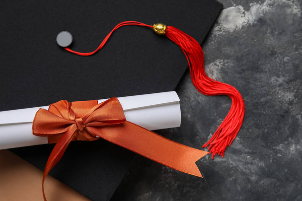 Diploma with red ribbon, graduation hat and book on dark grey table - Valokuva, kuva