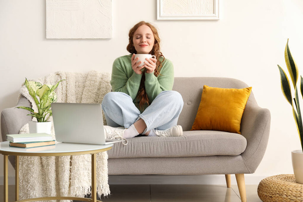 Beautiful redhead woman with cup of tea sitting on sofa at home - Zdjęcie, obraz