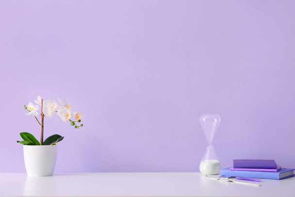 Posto di lavoro con fiore, clessidra, smartwatch e libri vicino alla parete lilla - Foto, immagini