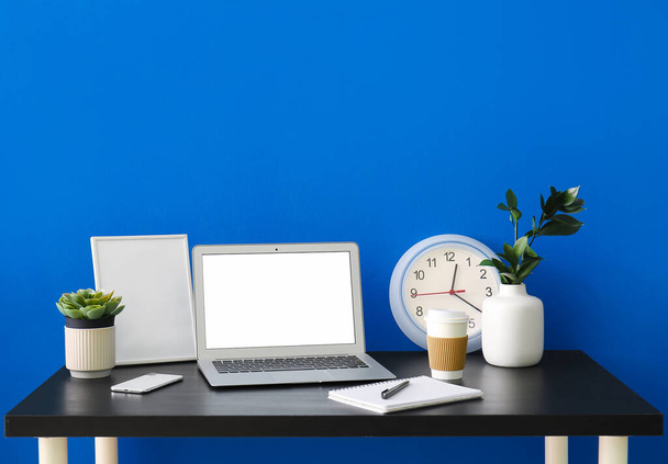 Luogo di lavoro con computer portatile, telaio, tazza di caffè e orologio vicino alla parete blu - Foto, immagini