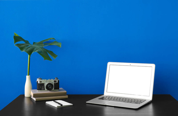 Χώρος εργασίας με φορητό υπολογιστή, φωτογραφική μηχανή και φύλλο παλάμης κοντά σε μπλε τοίχο - Φωτογραφία, εικόνα