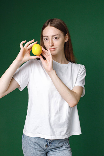 Indoor portrait of caucasian girl isolated on green background. Portrait of young beautiful woman in white t-shirt posing for a photo. Studio photo of a young girl holding a lemon. - Valokuva, kuva