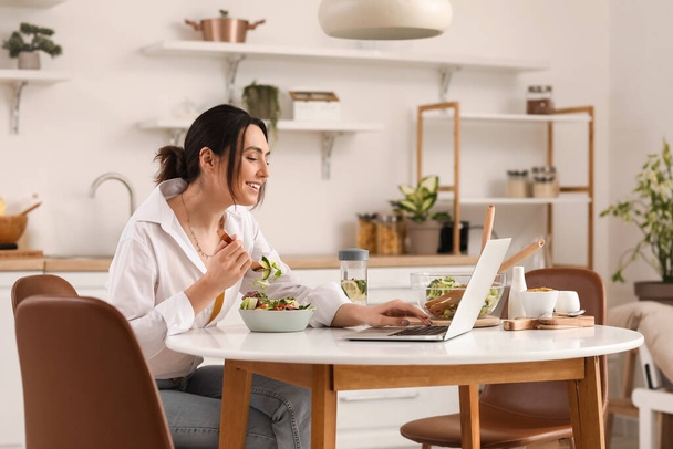 Jonge vrouw met laptop eten groentesalade aan tafel in de keuken - Foto, afbeelding