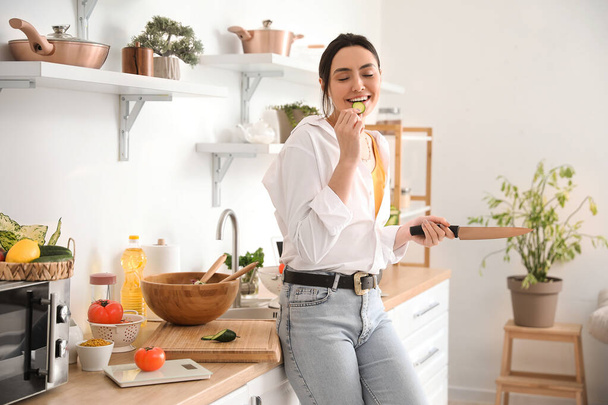 Mujer joven comiendo rodaja de pepino en la cocina - Foto, Imagen