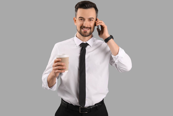 Hombre de negocios guapo con taza de café hablando por teléfono móvil sobre fondo gris - Foto, Imagen