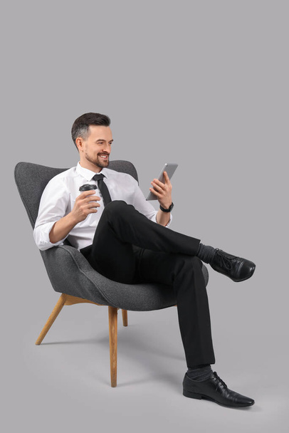 Jóképű üzletember tabletta számítógép és csésze kávé fotelben szürke háttér - Fotó, kép