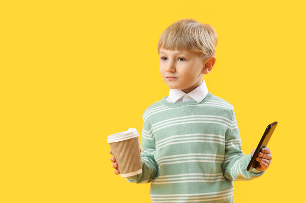 黄色の背景にコーヒーと携帯電話のカップを持つ面白い小さなビジネスマン - 写真・画像