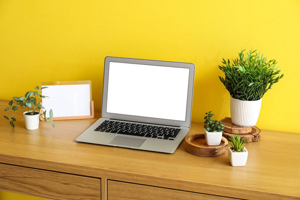 Рабочее место с искусственными растениями и ноутбуком возле желтой стены - Фото, изображение