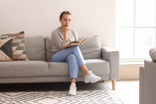 Átgondolt fiatal nő könyv ül kanapén otthon - Fotó, kép