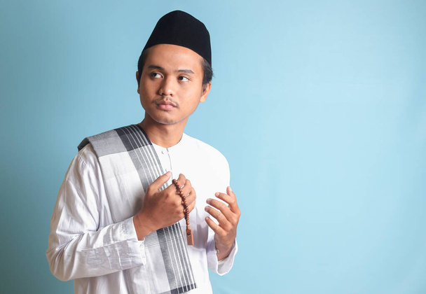 Ritratto di uomo musulmano asiatico in camicia koko bianca con copricapo che prega con le mani alzate. Immagine isolata su sfondo blu - Foto, immagini