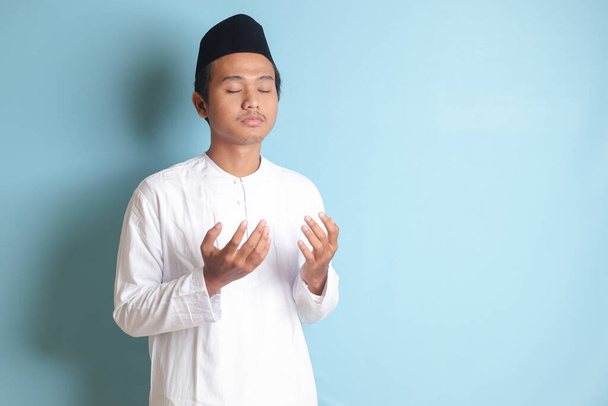 Portrét asijského muslima v bílém koko tričku s lebkovou čepicí, který se vážně modlí se zdviženýma rukama. Izolovaný obrázek na modrém pozadí - Fotografie, Obrázek