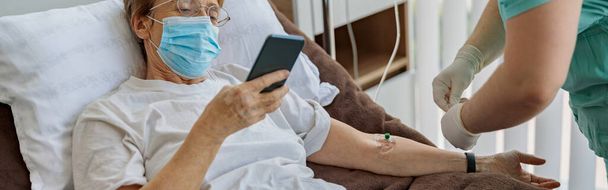 El paciente mayor enfermo usa el teléfono durante el procedimiento médico en la sala del hospital. Foto de alta calidad - Foto, imagen