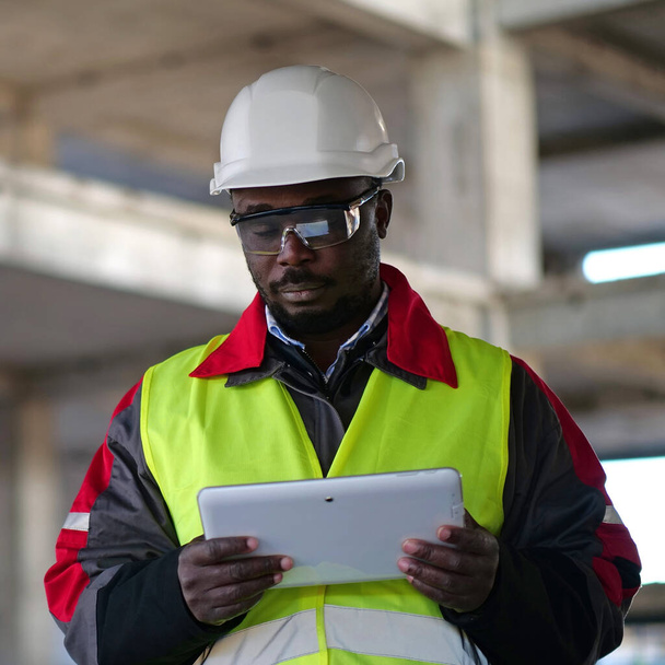 Afrykański amerykański pracownik w twardej czapce i okularach ochronnych z zamkniętymi oczami stoi na placu budowy, trzyma w ręku tablet - Zdjęcie, obraz