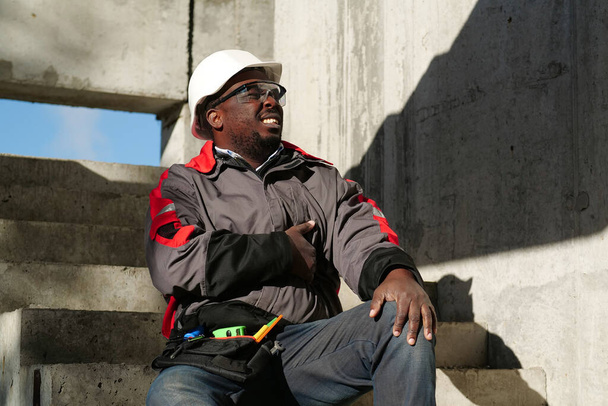 Αφροαμερικάνος εργάτης κάθεται στις σκάλες στο εργοτάξιο. Δεν αισθάνεται καλά και έχει στομαχόπονο, αγγίζει το στομάχι του. - Φωτογραφία, εικόνα