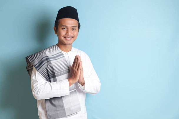 謝罪と歓迎の手のジェスチャーを示すskullcapと白いココシャツのアジアのイスラム教徒の男の肖像画。青い背景に孤立したイメージ - 写真・画像