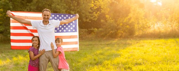 Vlastenecká dovolená. Šťastná rodina, matka a dcery s americkou vlajkou venku při západu slunce. USA slaví Den nezávislosti 4. července - Fotografie, Obrázek