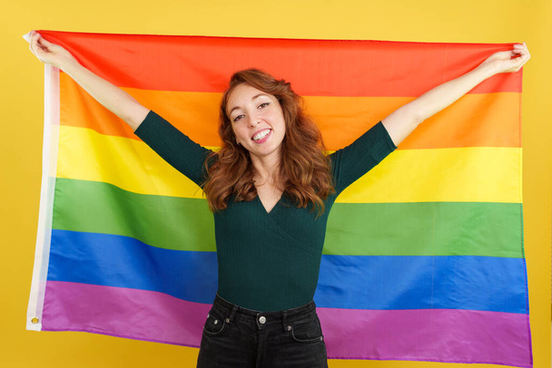 gelukkig roodharige vrouw het verhogen van een lgbt regenboog vlag in studio met gele achtergrond - Foto, afbeelding