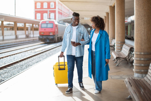 訓練に行く。鉄道駅でスーツケースとハッピーブラックカップルウォーキング,荷物とチャットを運ぶ陽気な若いアフリカ系アメリカ人の配偶者,一緒に旅行を楽しむ,スペースをコピー - 写真・画像