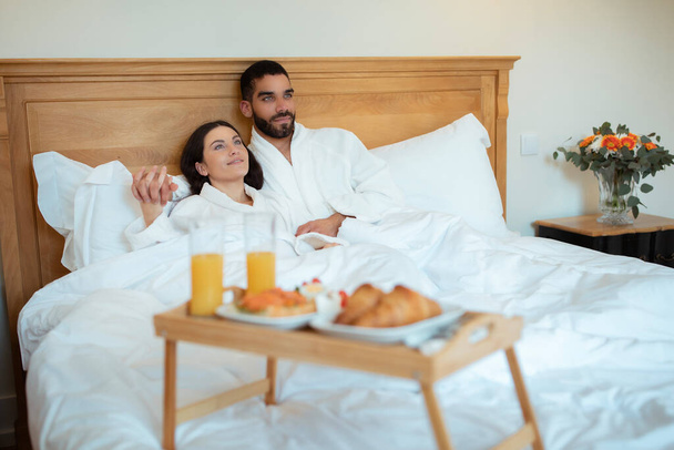 Loving para przytulanie Korzystanie śniadanie w łóżku Leżąc blisko taca stołowa z pysznym jedzeniem w nowoczesnym pokoju hotelowym w pomieszczeniach, nosząc szlafroki. Pojęcie wakacyjnego miesiąca miodowego. Skupienie selektywne - Zdjęcie, obraz
