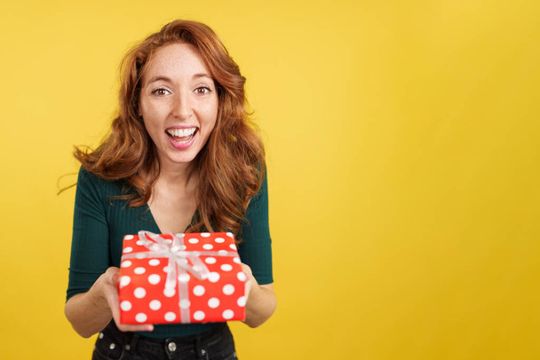 Redheaded woman smiling at camera with a gift in studio with yellow background - Valokuva, kuva