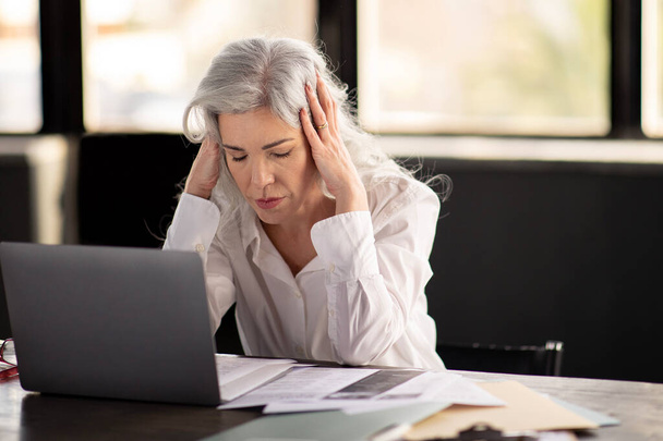 Migraine. Femme d'affaires mature souffrant de maux de tête douleur, en appuyant sur les mains à la tête douloureuse assis avec les yeux fermés à l'ordinateur portable travaillant en ligne dans le bureau à l'intérieur. Problèmes de santé - Photo, image