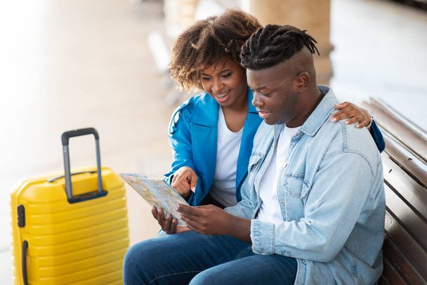 Allegri giovani sposi neri che controllano il percorso di viaggio con la mappa della città mentre siedono sul banco alla stazione ferroviaria, felice coppia afro-americana che sceglie la destinazione di viaggio insieme in attesa di treno - Foto, immagini