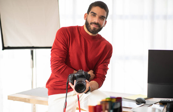 Positiv lächelnder hübscher hispanischer junger Mann in stylischem lässigem Outfit, professioneller Fotograf posiert in seinem Fotostudio am Arbeitsplatz, sitzt am Schreibtisch mit PC, hält Digitalkamera, Kopierraum - Foto, Bild