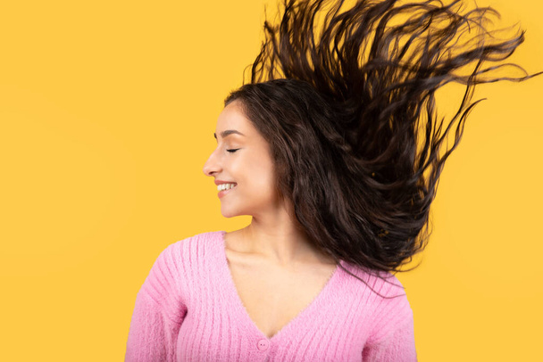 Satisfied millennial middle eastern female brunette shaking her head with long hair, has fun, enjoy lifestyle, freedom, isolated on yellow studio background. Relax, people emotions - Photo, image