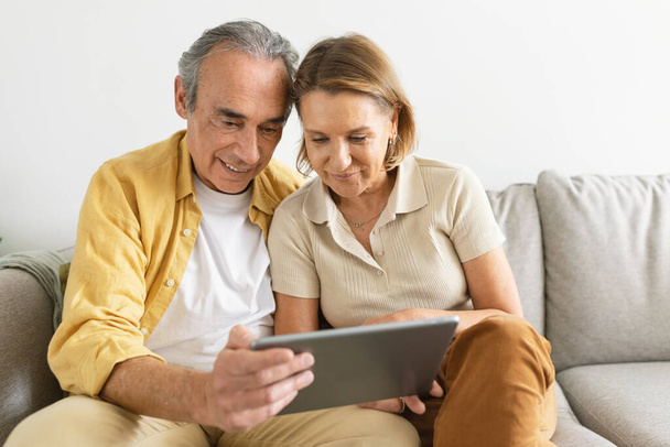 Hymyilevä vanhempi pari, joka käyttää taulutietokonetta, surffaa internetissä, katsoo elokuvaa verkossa yhdessä, istuu sohvalla kotona. Ikääntyneet puolisot käyttävät digitaalista gadgetia viikonloppuna - Valokuva, kuva