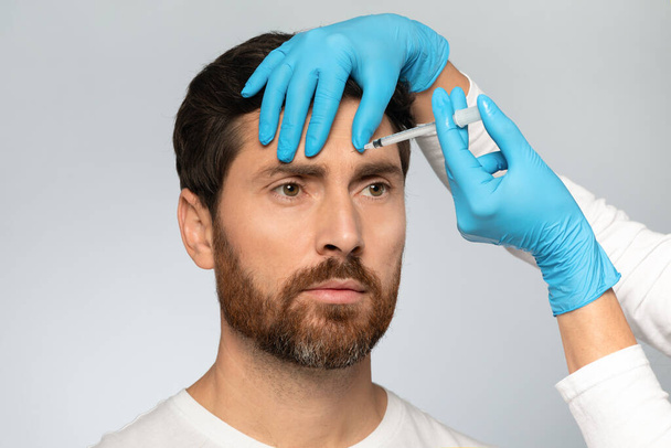 Handsome middle aged bearded man getting brow lift, beautician in gloves making beauty injection for handsome male, standing over grey background - Zdjęcie, obraz