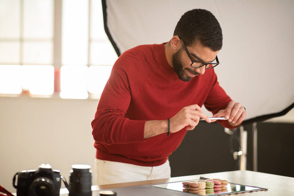 Handsome young hispanic bearded man wearing casual outfit and eyewear photographing delicious tasty sweet dessert colorful macaroons on mirror board, using smartphone, photo studio interior - Photo, Image