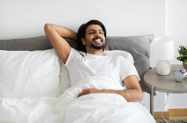 Onnellinen komea nuori Lähi-idän mies valkoisessa t-paidassa makaa sängyllä, haaveilee, nauttii vapaa-ajasta, lepää, rentoutuu makuuhuoneen sisustuksessa, kopioi tilaa. Viikonloppu yksin ja hyvää huomenta kotona - Valokuva, kuva