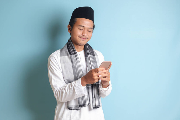 Portrait of young Asian muslim man holding and touching mobile phone with smiling expression on face. Isolated image on blue background - Фото, изображение