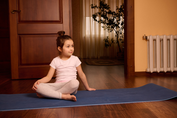 Приємна чудова кавказька 5-річна дівчинка в активному одязі, посміхається, озирається, сидить на комірці йоги в затишному будинку. Щасливі діти. Дитинство. Спорт, спосіб життя та хобі. - Фото, зображення