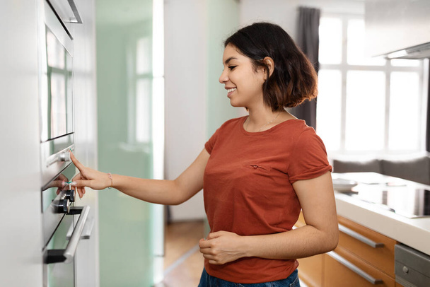 Happy Young Arab Housewife Using Modern Stove While Preparing Food At Home, Smiling Middle Eastern Woman Turning On Electric Oven In Kitchen Interior, Pressing Button With Finger, Copy Space - Фото, зображення