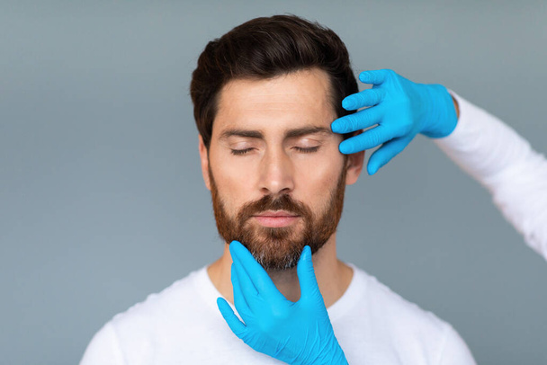 Portret brodatego mężczyzny w średnim wieku przygotowującego się do operacji plastycznych, lekarz dotykający męskiej skóry twarzy, wyizolowany na szarym tle, przestrzeń do kopiowania - Zdjęcie, obraz