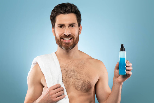 Όμορφος μεσήλικας άνδρας με πετσέτα στον ώμο που δείχνει μπουκάλι με μπλε καλλυντικό υγρό, ποζάροντας πάνω από το μπλε φόντο στούντιο και χαμογελώντας στην κάμερα, ελεύθερος χώρος - Φωτογραφία, εικόνα