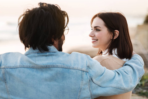 Smiling millennial indian guy hugging caucasian woman, enjoy romantic and love at free time on beach near ocean. Relax and rest together, picnic, vacation, date in nature, close up - Photo, Image