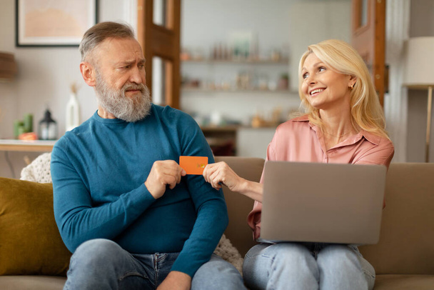 Happy Mature Shopaholic Wife Shopping Online On Laptop Taking Credit Card From Displeased Greedy Husband Sitting On Sofa At Home. Ecommerce And Internet Banking Service Advertisement - Φωτογραφία, εικόνα