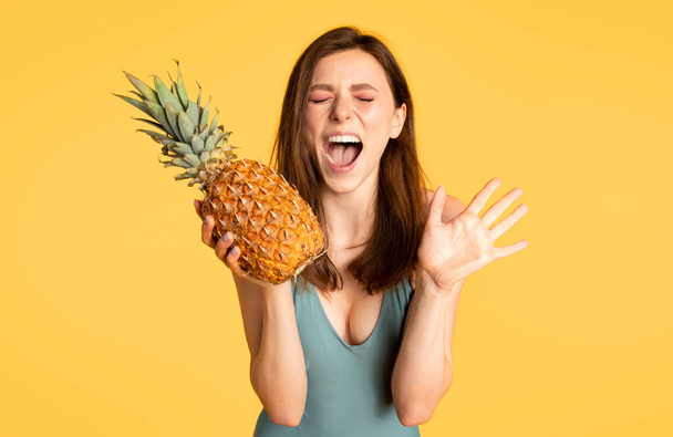 Excited woman in swimsuit holding beautiful and juicy ripe pinapple and shouting, young lady enjoying summer vacation, standing on yellow background, copy space - Photo, Image