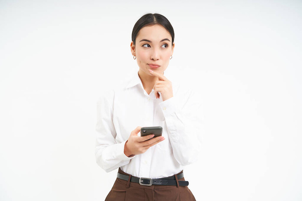 Azjatycka kobieta biznesu, młody menedżer trzyma smartfona i myśli, wygląda troskliwie na bok, podejmuje decyzje, stoi na białym tle. - Zdjęcie, obraz