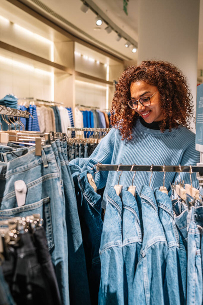 Αφρικανική αμερικανική νεαρή γυναίκα με σγουρά μαλλιά σε casual ρούχα επιλέγοντας τζιν, ενώ επισκέπτονται εμπορικό κέντρο - Φωτογραφία, εικόνα