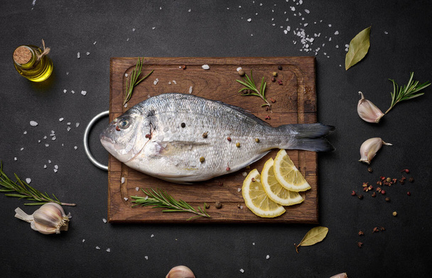 Сырая целая рыба Дорадо на деревянной кухонной доске и специи для приготовления пищи, вид сверху на черный стол - Фото, изображение