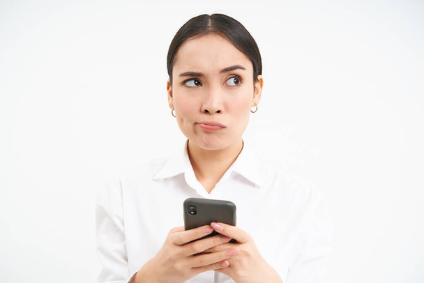 Obrázek asijské prodavačky, office manager drží smartphone, pracuje na textu pro sociální média, pomocí mobilu, bílé pozadí. - Fotografie, Obrázek
